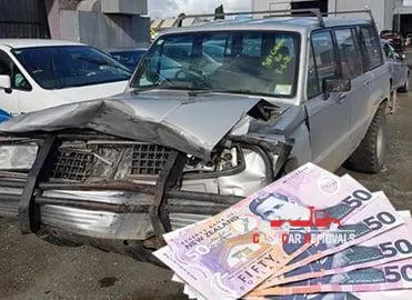 A heavily damaged silver SUV in a wrecking yard with a fan of New Zealand fifty-dollar cash notes in the foreground, representing cash for cars South Auckland services.